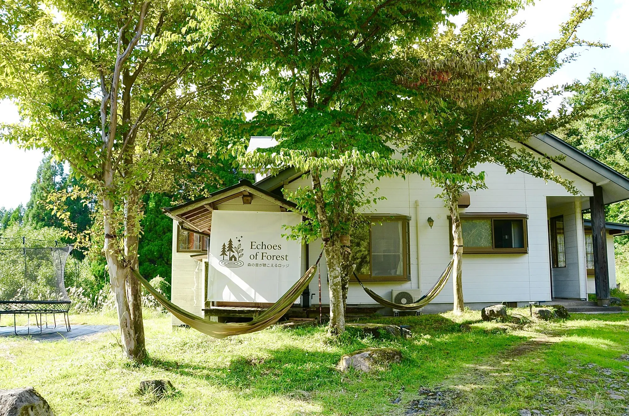 The white timber exterior of Echoes of Forest lodge in Minamioguni in summer, with hammocks strung between trees on the lawn