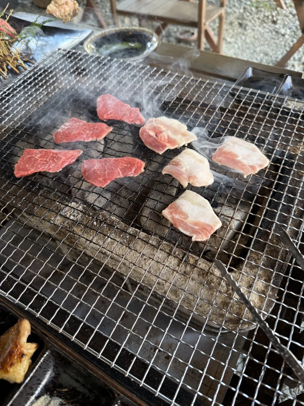 Slices of akaushi beef and pork belly grilling on a wire mesh over glowing charcoal, smoke rising