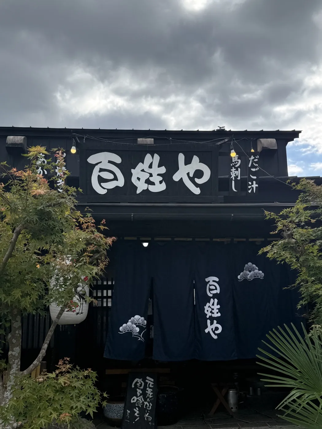 The entrance of Hyakushoya in Oguni: a black timber building with the name in large white characters, navy noren curtains with a pine motif, and hanging signs for akaushi, horse sashimi and dago-jiru