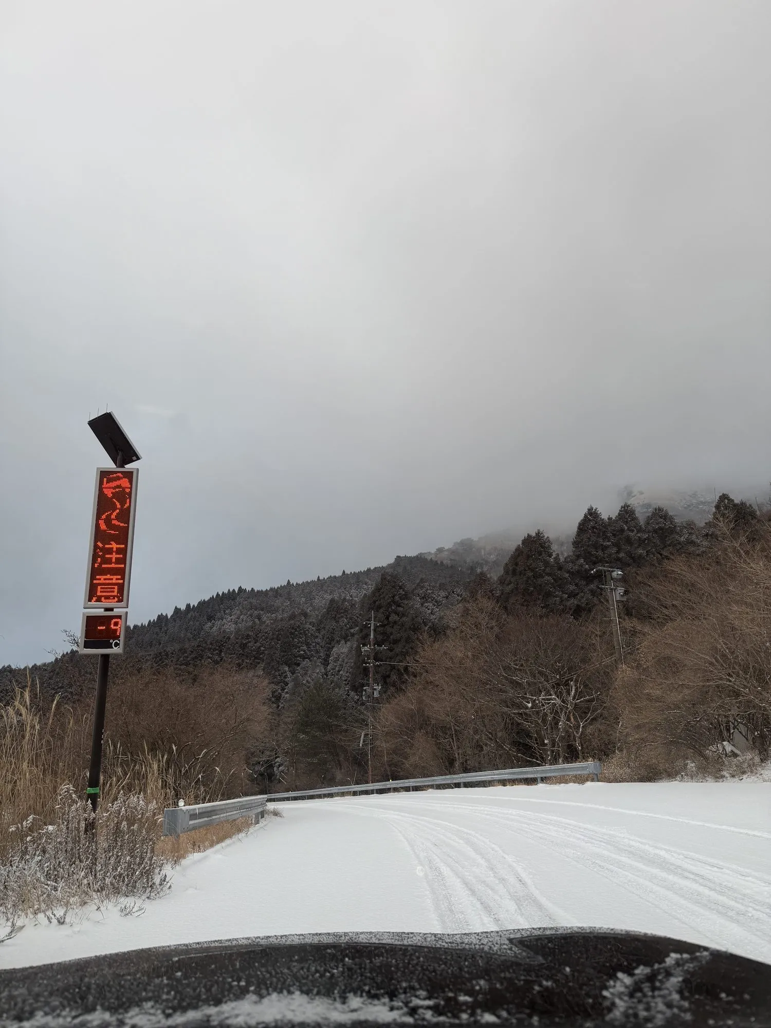 An electronic roadside sign near the Kuju ski area reading 凍結注意 — "caution, ice" — above a temperature readout showing minus 9°C, with snow-covered road ahead and cloud sitting on the ridge