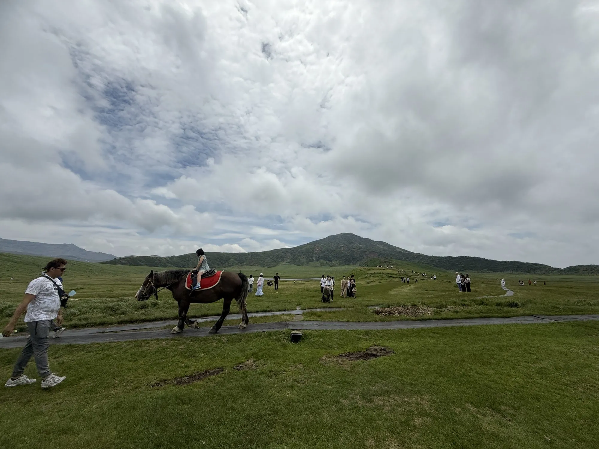 Horses and walkers on the green grassland plain of Kusasenri at Aso, with a volcanic cone behind under heavy cloud