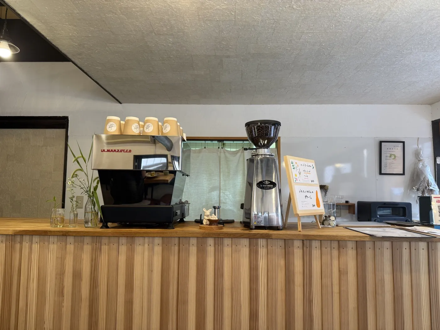 The cedar counter at SLOW WALK CAFE with a La Marzocco espresso machine, grinder and a row of takeaway cups, against a white wall