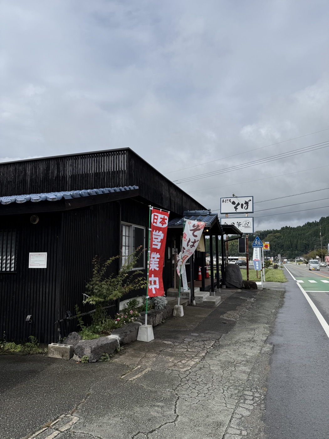 黒い板張りの建物に「本日営業中」ののぼり。国道沿いに「そば」の看板が立つ